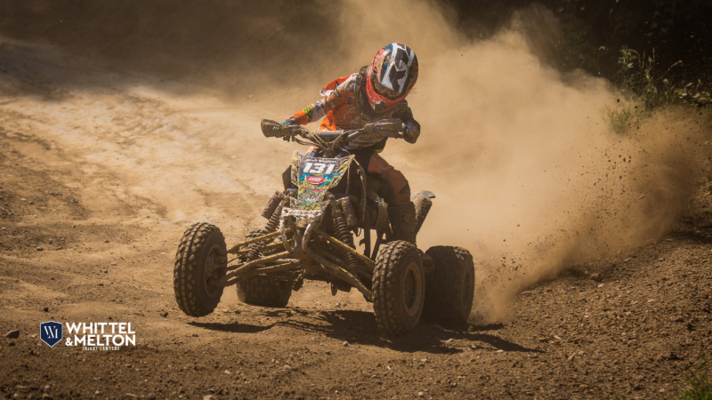 Child riding an ATV on a dirt track, kicking up dust, wearing a helmet and safety gear.

