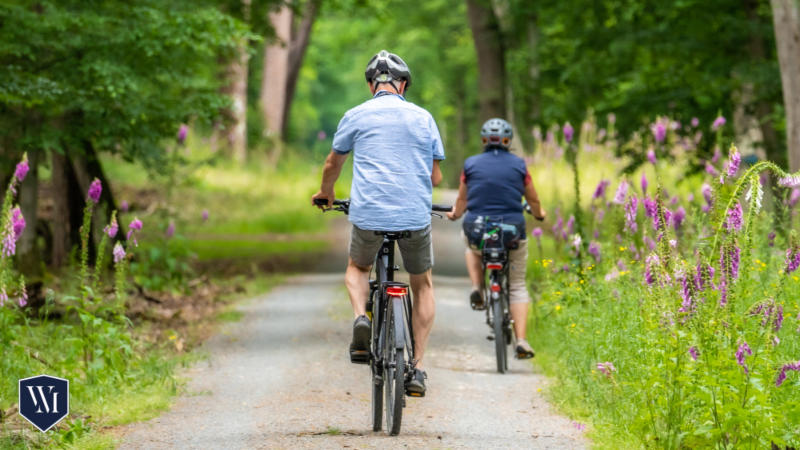 Two cyclists wearing helmets ride along a scenic tree-lined path surrounded by blooming wildflowers, emphasizing safe and enjoyable biking in nature. A Whittel & Melton logo is visible in the bottom left corner.